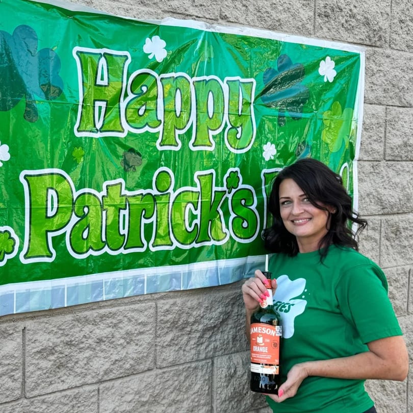 Woman in a green Wolfies shirt holding a Jameson Orange bottle in front of a "Happy Patrick's Day" banner.