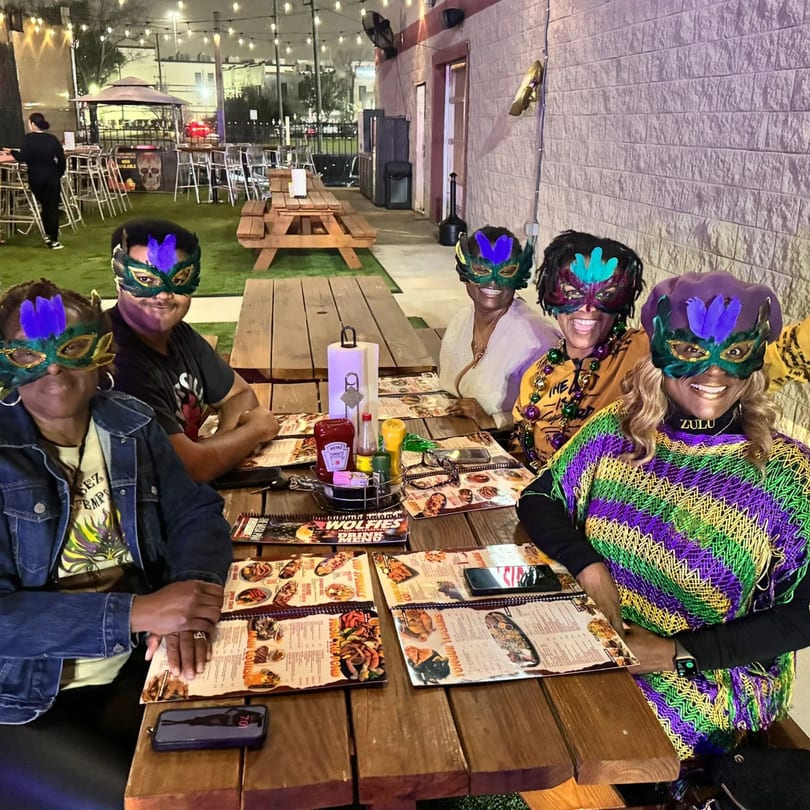 Five people in Mardi Gras masks and attire sit at a picnic table on Wolfies Restaurant patio.