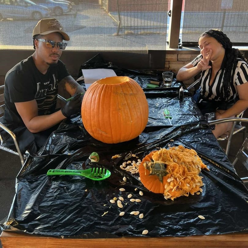 Two people carving pumpkins at a table covered with a black tarp at Wolfies Sports Bar.