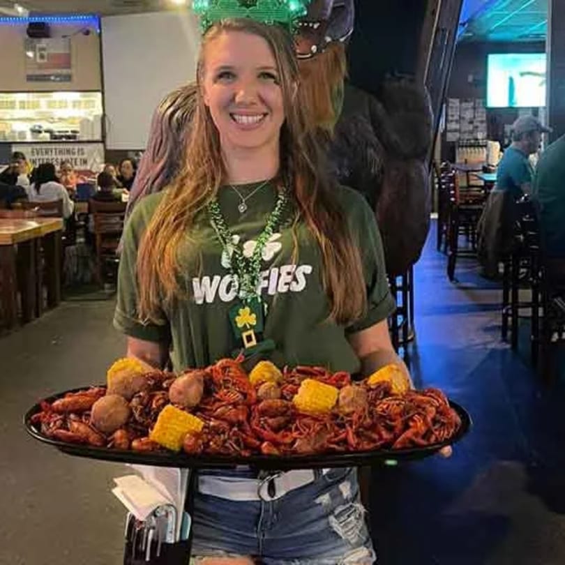 A smiling Wolfies server holds a large tray of crawfish, corn, and potatoes in a lively sports bar.