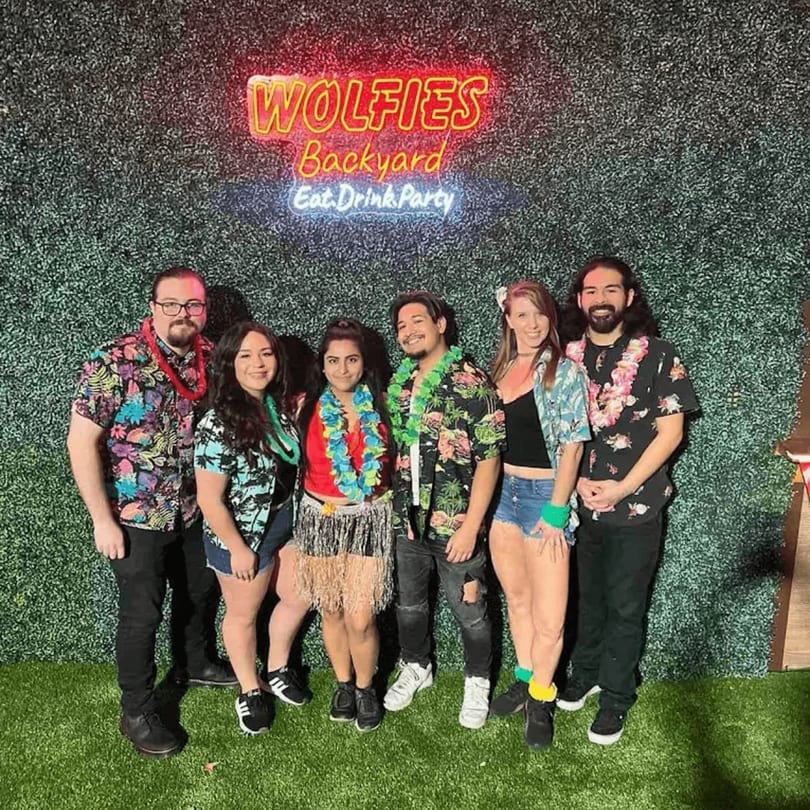 Six people in Hawaiian shirts and leis pose in front of a Wolfies Backyard neon sign.