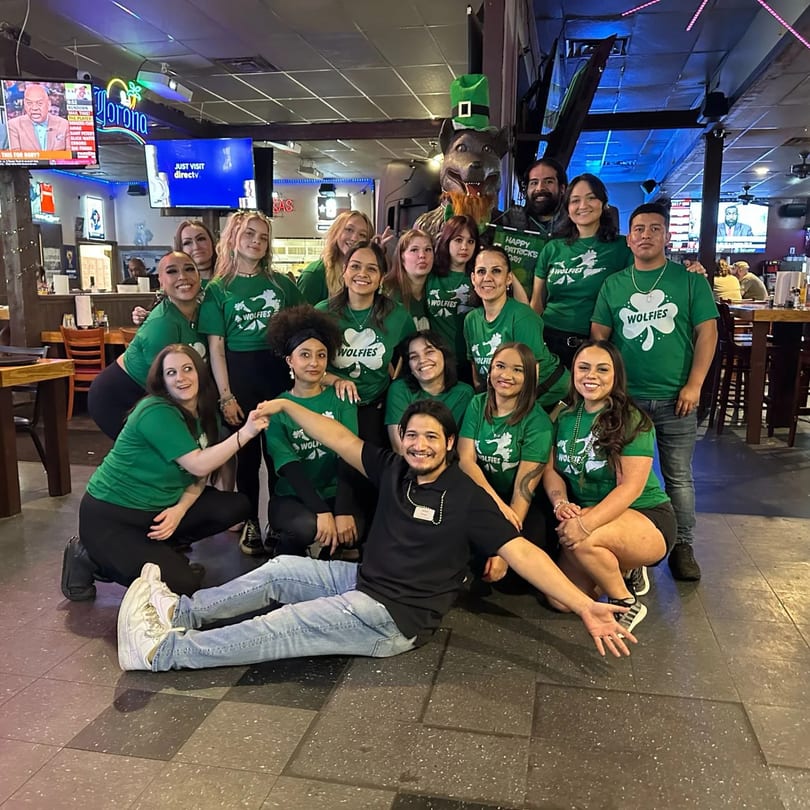 Wolfies Restaurant staff in green shirts with shamrocks, celebrating St. Patrick's Day in a sports bar.