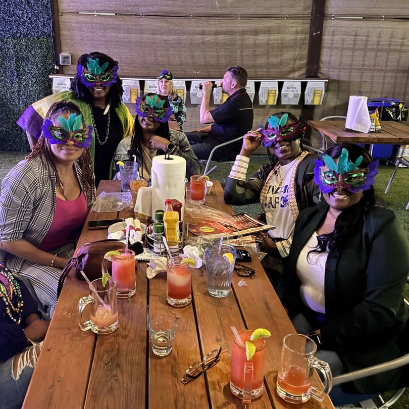 Five women in colorful Mardi Gras masks and a man drinking at a wooden table in a sports bar.