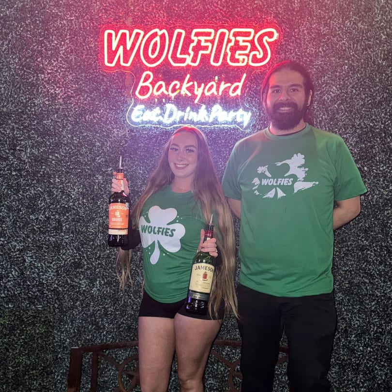 Two Wolfies staff members in green shirts holding Jameson bottles under a neon sign.